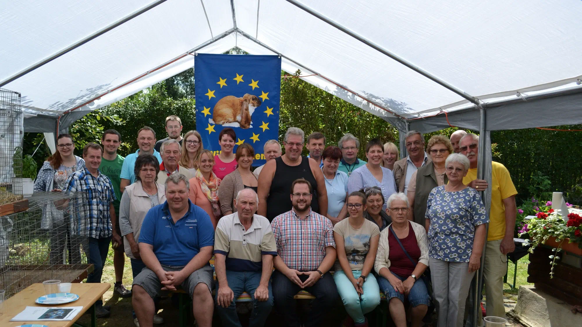Photo de groupe des participants à la rencontre de l'IG 2017 sous une tente blanche, au milieu un drapeau bleu avec une couronne d'étoiles et l'image d'un lapin