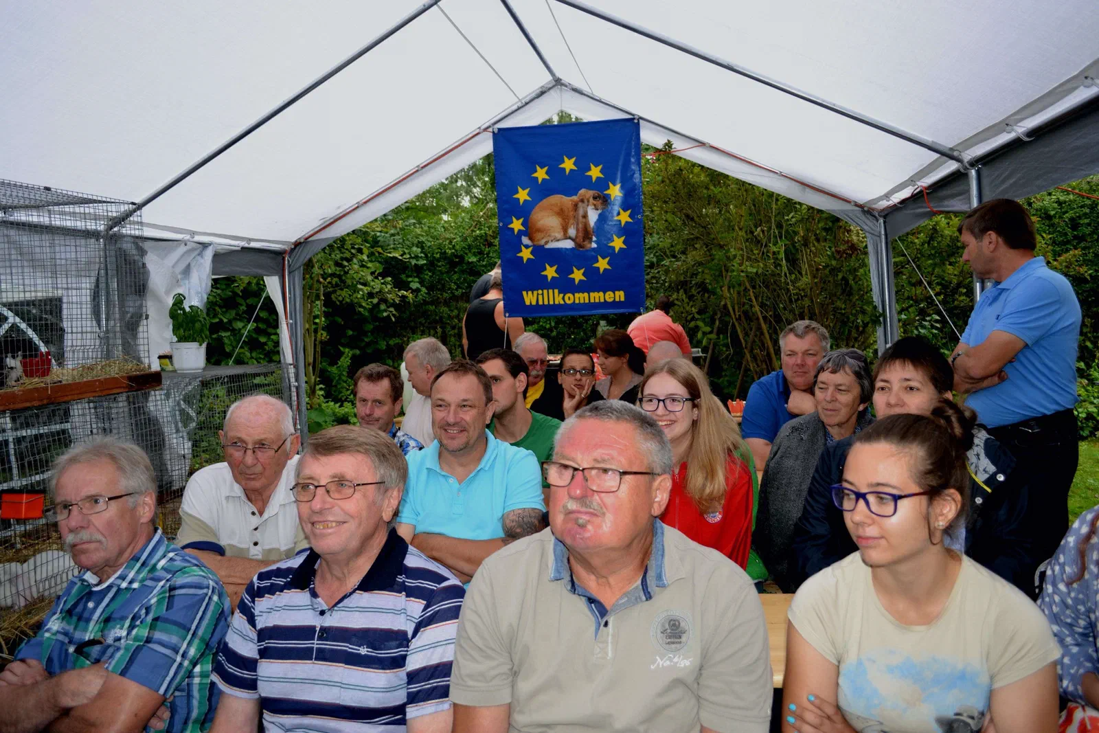 Invités et juges assis sur des bancs sous la tente, derrière eux un drapeau bleu avec l&rsquo;image d&rsquo;un lapin et l&rsquo;inscription « Willkommen » (bienvenue)