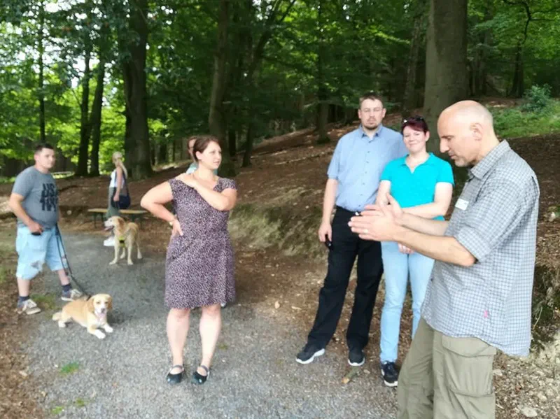 En museumsguide forklarer noget for en lille gruppe deltagere på en skovsti, i baggrunden to hunde