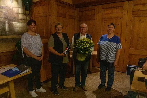 Fire personer står ved siden av hverandre i et trepanelt rom, en mann holder en potte med gule blomster, en kvinne en flaske vin