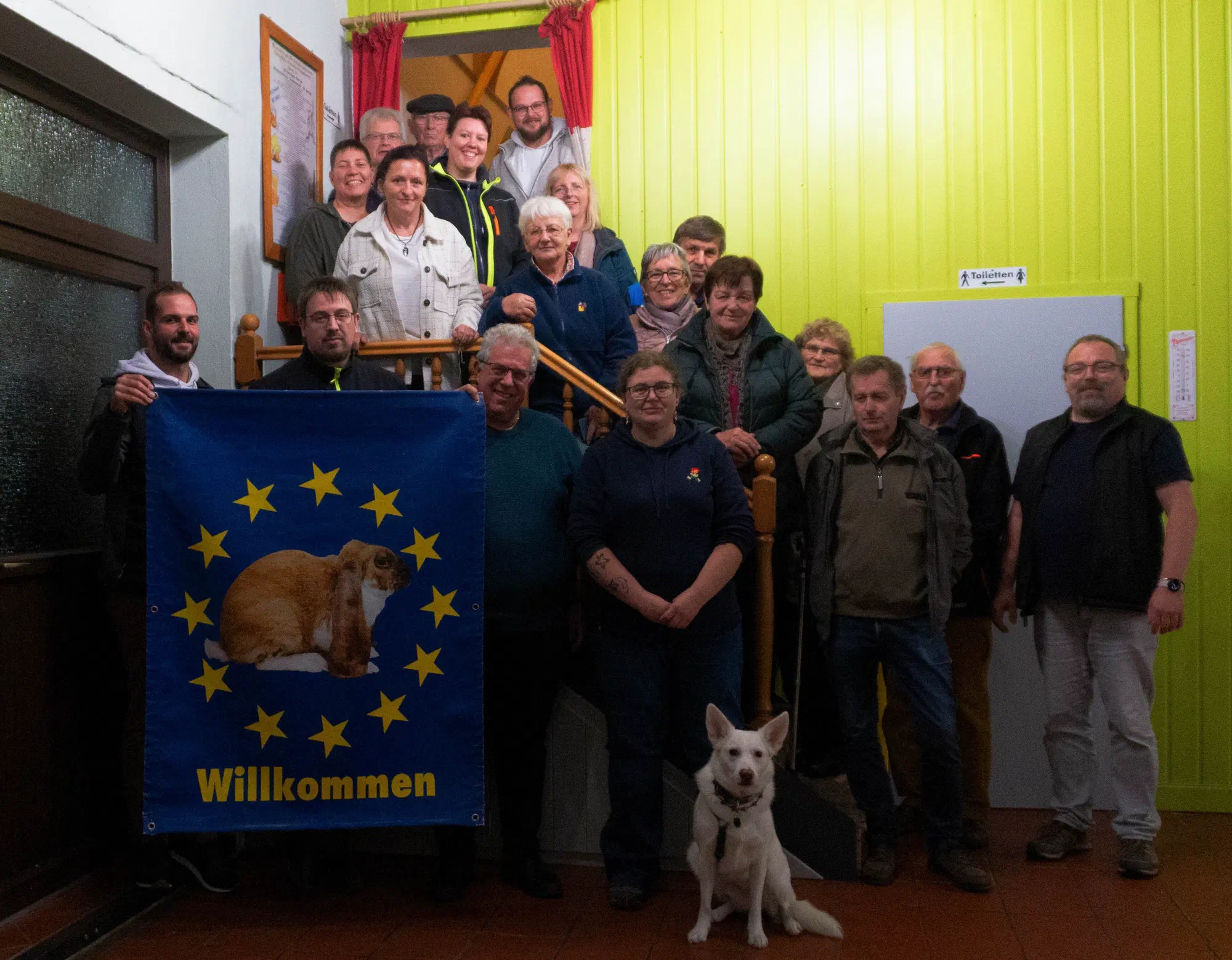 Photo de groupe des participants dans une cage d'escalier à la paroi en bois vert-jaune, devant un drapeau bleu avec un lapin et l'inscription Willkommen (bienvenue) et un chien blanc